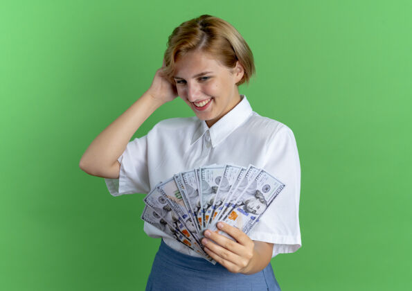 俄罗斯人年轻微笑的金发俄罗斯女孩抱着钱看着手放在头上孤立的绿色背景和复制空间钱年轻手