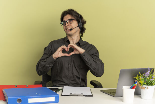 手势戴着耳机 戴着眼镜的年轻上班族坐在办公桌旁 拿着办公工具 使用笔记本电脑 手势 心形手势 绿色背景和复印空间使用办公桌工人
