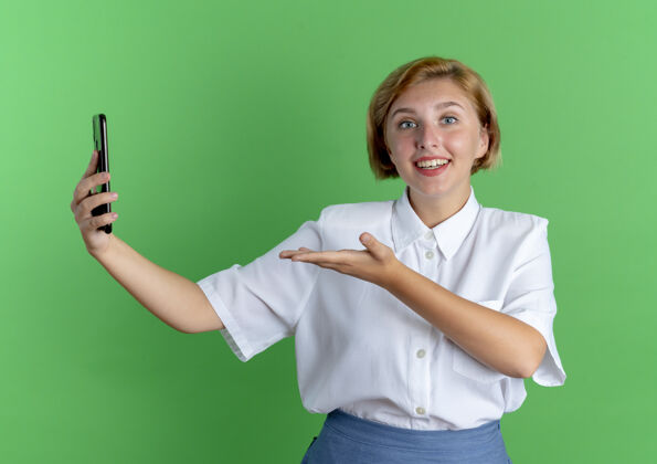 俄罗斯人年轻惊讶的金发俄罗斯女孩拿着和手指在绿色背景与复制空间孤立的电话金发电话年轻人