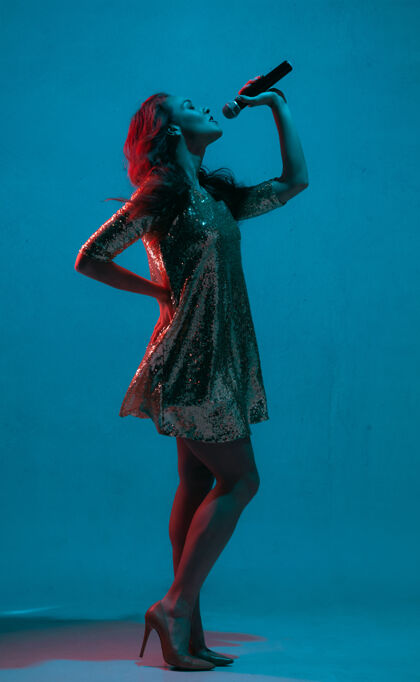 情感白种人女歌手的肖像被隔离在霓虹灯下的蓝色工作室背景上漂亮的女模特穿着明亮的裙子 带着麦克风人类情感的概念 面部表情 广告 音乐 艺术行政人员音乐家音乐
