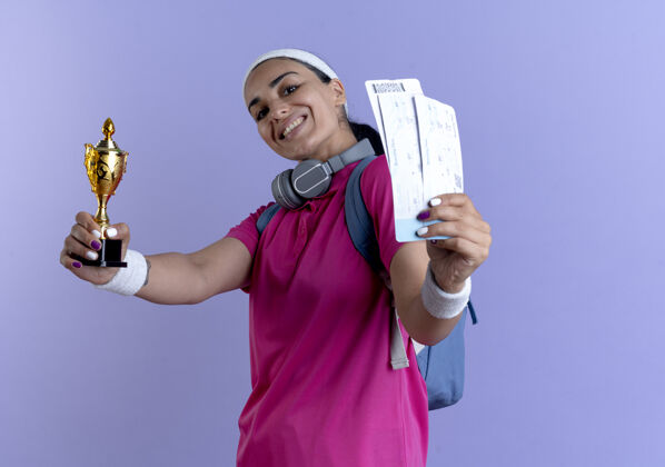 运动年轻的笑容可掬的白人运动女性 戴着背包头带和腕带 手持优胜者杯和机票 背景为紫色 留有复印空间包年轻微笑