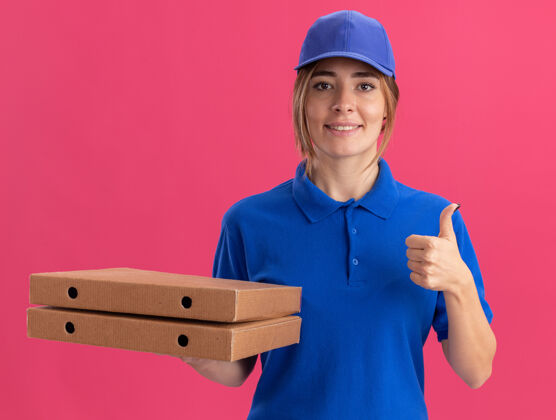 微笑身着制服 面带微笑的年轻漂亮女送货员竖起大拇指 把比萨饼盒孤立地放在粉红色的墙上披萨脸递送