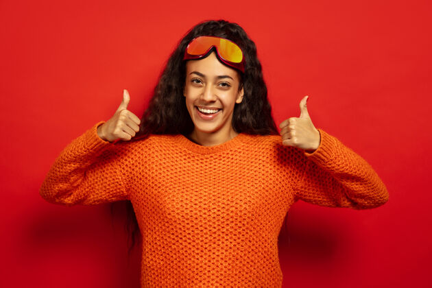 女性美国黑人年轻黑发女子的肖像滑雪面具在红色工作室的背景概念的人类情感 面部表情 销售 广告 冬季运动和假日微笑和拇指向上表情手女性