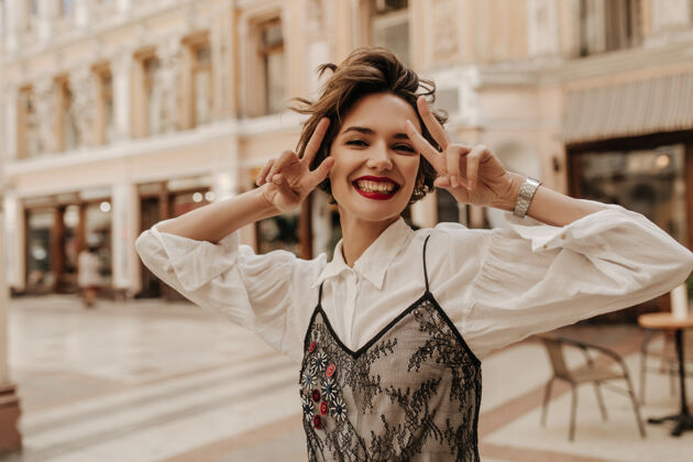 时尚快乐的红唇女人在城市里展示着和平的标志短发 白衬衫 黑蕾丝的现代女人在街上微笑着姿势女性短发