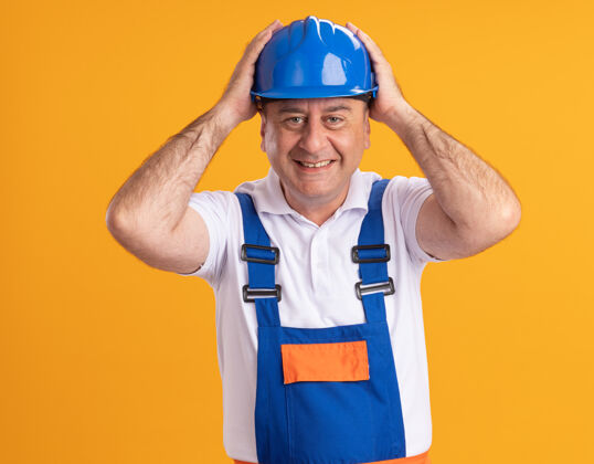 姿势身着制服的微笑的成年建筑工人把手放在隔离在橙色墙上的头上脸感觉人
