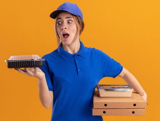 制服焦急的年轻漂亮的女送货员穿着制服拿着纸食品包装和比萨饼盒上的容器隔离在橙色的墙上人焦虑纸