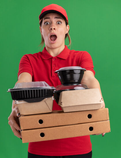 站着焦急的年轻漂亮的女送货员穿着制服拿着纸食品包装和比萨饼盒上的容器隔离在绿色的墙壁上披萨食物漂亮