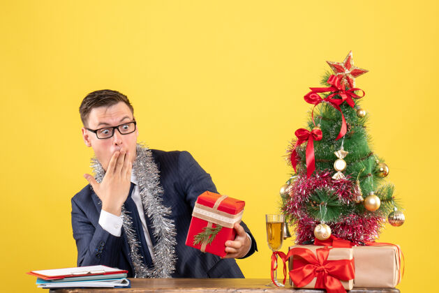 树前视图惊讶的男人看着他的礼物坐在圣诞树旁边的桌子上 黄色背景上的礼物附近桌子礼物