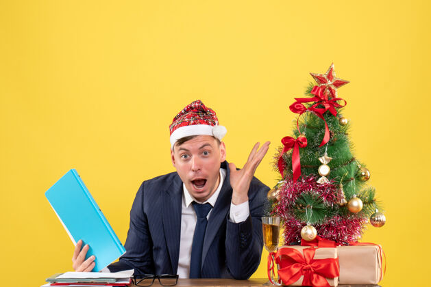 人前视图惊奇的商人坐在圣诞树旁的桌子上 呈现黄色背景工作前面背景