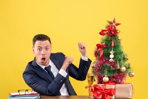 人正面图惊奇的男人手指着后面坐在圣诞树旁的桌子和黄色背景上的礼物礼物前面办公室