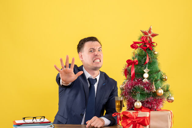 背景前视图愤怒的男人在圣诞树旁的桌子旁做停车标志 黄色背景上的礼物树前面男