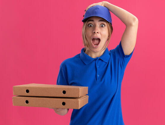 年轻人穿着制服的年轻漂亮的送货员兴奋地把手放在帽子上 手里拿着隔离在粉红色墙上的比萨饼盒姿势女孩站着