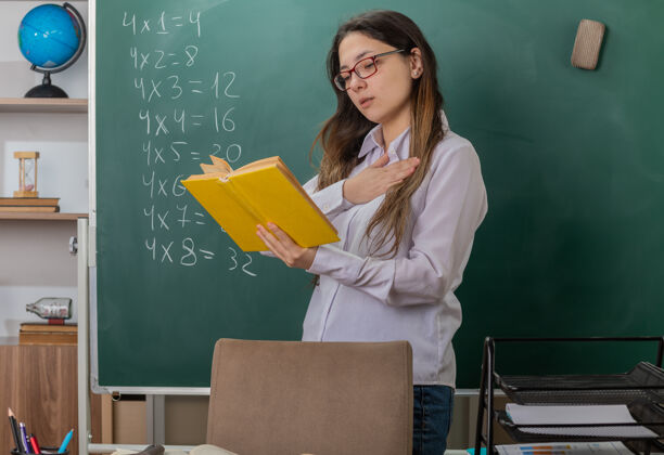 桌子年轻的女老师戴着眼镜看书讲解课 手放在胸前 自信地站在教室黑板前的课桌旁解释学校年轻