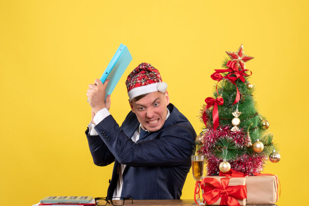 树前视图愤怒的商人将文件从圣诞树旁的桌子上扔掉 并在黄色背景上呈现工作肖像附近
