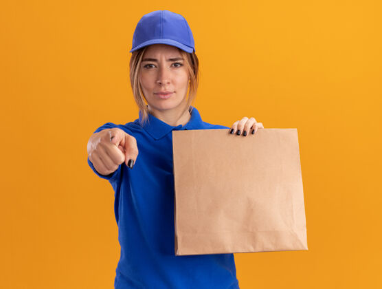 姿势自信的年轻漂亮的女送货员穿着制服 手里拿着纸包 指着橘黄色的墙上孤立的前面交货制服公民