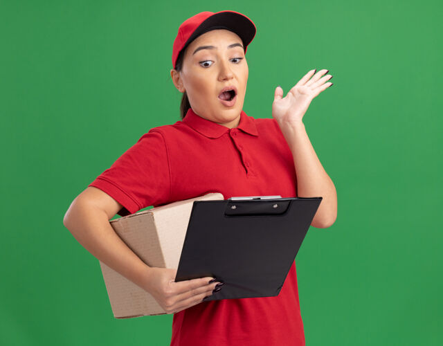 制服年轻的送货员身穿红色制服 头戴鸭舌帽 手里拿着一个纸板箱 手里拿着剪贴板 站在绿色的墙上 惊讶地抬起胳膊看着它站着纸板胳膊