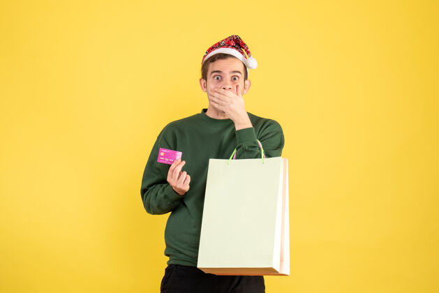 人正面图：戴着圣诞帽的年轻人拿着购物袋和卡片 手放在黄色背景上的嘴巴上秘书圣诞节举行