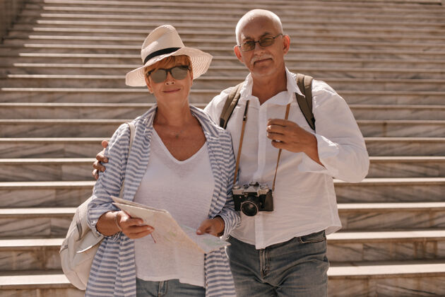 墙戴着帽子 戴着墨镜 穿着条纹衬衫的女人拿着卡片 和留着胡子 穿着白衬衫的男人用相机拥抱女士外面游客