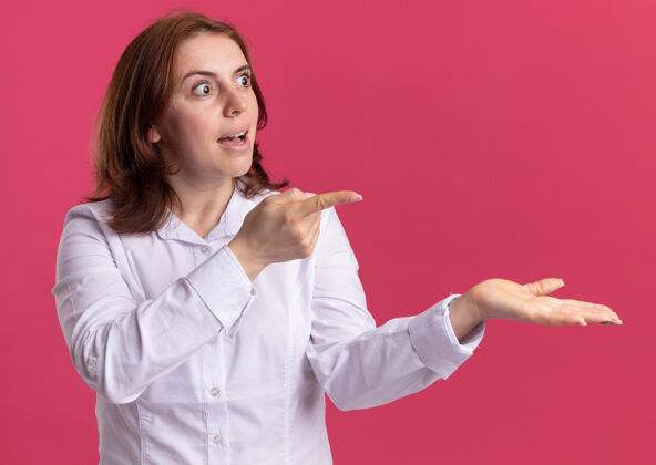 目录穿白衬衫的年轻女子一边看一边困惑地用食指指着呈现的东西 胳膊站在粉红色的墙上手臂手指立场