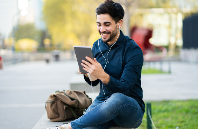 男性年轻人坐在户外的长椅上通过数码平板电脑进行视频通话的照片城市概念触摸屏城市休闲