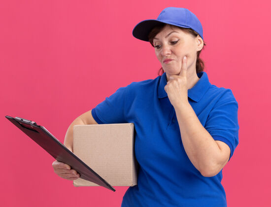 剪贴板身穿蓝色制服 头戴鸭舌帽的中年送货妇女站在粉红色的墙上 严肃地看着剪贴板纸板严重中年