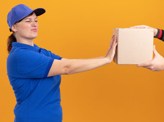 送货身穿蓝色制服 戴着帽子的年轻送货员站在橘色的墙上 表情厌恶 满脸忧虑 拒绝拿包裹包装帽子站着