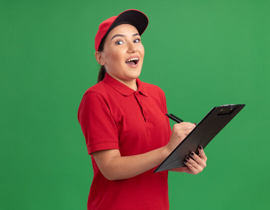 剪贴板年轻的送货员身穿红色制服 戴着帽子 拿着写字板和铅笔 站在绿色的墙上 高兴而兴奋地看着前面快乐制服铅笔