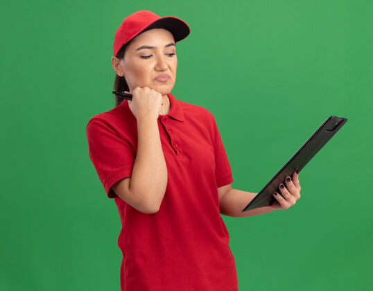 交货年轻的女送货员穿着红色制服 戴着帽子 拿着剪贴板 站在绿色的墙上困惑地看着它拼图女人年轻