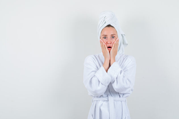 惊喜照片中的年轻女子手放在脸颊上 穿着白色浴衣 拿着毛巾 脸上露出惊讶的表情人健康积极