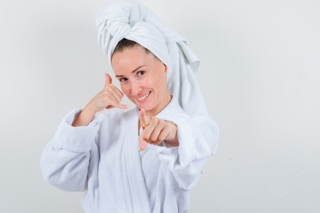 时尚年轻女子展示手机手势 指着相机 穿着白色浴衣 毛巾 看上去很开心 正对着镜头女性表演在一起