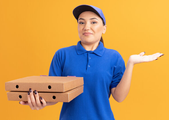 年轻身穿蓝色制服 戴着帽子的年轻送货员拿着披萨盒 面带微笑困惑地站在橘色的墙上 抬起手臂抱着站着女人