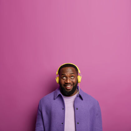 音乐工作室拍摄的快乐未剃胡子的男人戴着耳机听音频 微笑着看向上方 穿着紫色衬衫 拥有健康的深色皮肤美国非洲空白