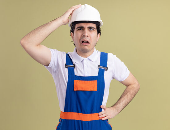 年轻年轻的建筑工人穿着建筑制服 戴着安全帽 困惑地看着前面 手放在头上 困惑地 非常焦虑地站在绿色的墙上困惑头建设者