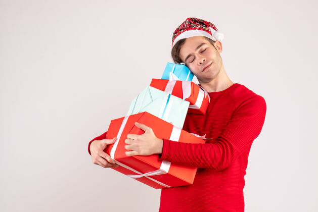 关闭正面视图闭着眼睛的年轻人戴着圣诞帽紧紧地拿着他的礼物在白色背景上复制空间眼睛微笑帽子