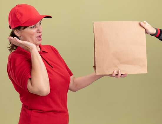 感受身穿红色制服 戴着帽子的年轻送货员站在绿色的墙上 一边接受纸包裹 一边兴奋不已送货女人帽子