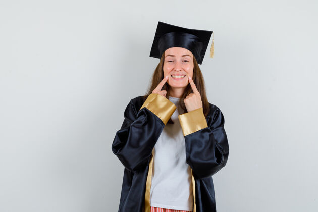 学位女毕业生指着她穿着学院服的牙齿 看上去很高兴正面图长袍微笑大学