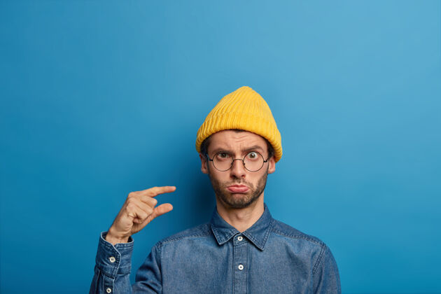 手指悲伤不高兴的男人的照片塑造小物体 演示小项目 收到不多 穿着时髦的黄色帽子和牛仔衬衫手势可怜帽子