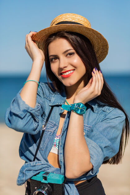 脸微笑的女人戴着草帽 穿着时髦的夏装 在沙滩上与复古相机合影娱乐享受乐趣