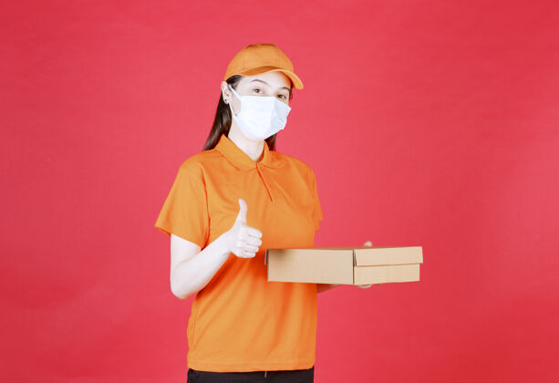 成人身着橙色制服 戴着面具的女快递员手持一个纸板箱 手上有明显的手势订单口罩服务