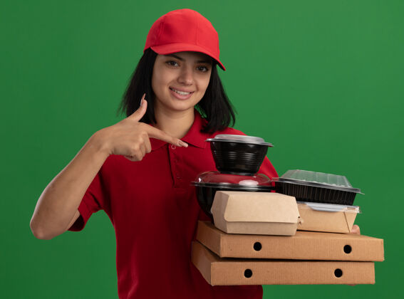 送货身着红色制服 头戴鸭舌帽 手持比萨饼盒和食品包的快乐年轻送货女孩站在绿色的墙上微笑着指着他们手指食物女孩