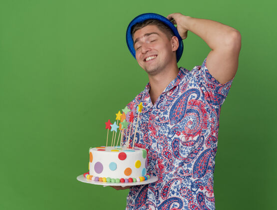 小伙子笑容满面的年轻人闭着眼睛戴着蓝色帽子拿着蛋糕把手放在脑后绿色背景上帽子手年轻人