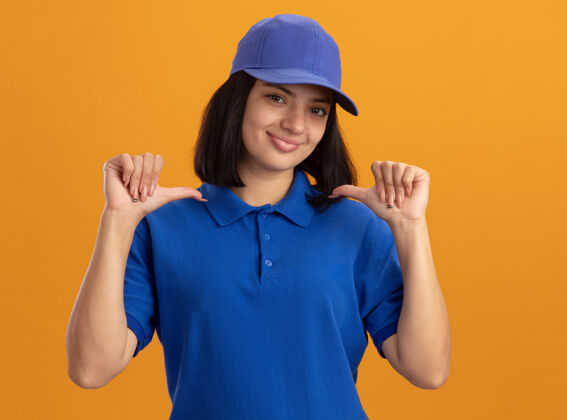 帽子身穿蓝色制服 头戴鸭舌帽的年轻送货员自信地微笑着指着站在橙色墙上的自己他自己微笑自信