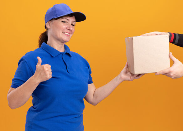站立身着蓝色制服 头戴鸭舌帽的年轻女送货员站在橙色的墙上 友好地展示着收包裹的ok标志微笑Ok制服