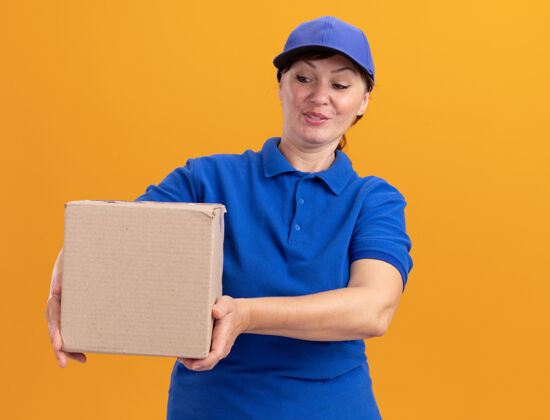 纸板快乐的中年送货妇女 身穿蓝色制服 戴着帽子 手里拿着纸板箱 站在橙色的墙上微笑着看着它制服送货盒子