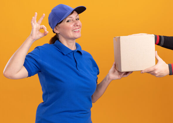 盒子身着蓝色制服 头戴鸭舌帽的中年女送货员向站在橙色墙上的顾客微笑友好地展示ok标志表演中间顾客