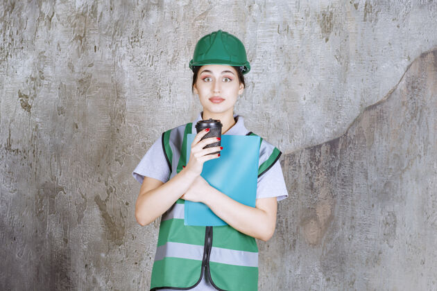 制服身着绿色制服 头戴安全帽的女工程师 手里拿着一个黑咖啡杯和一个蓝色项目文件夹检查表职员工人