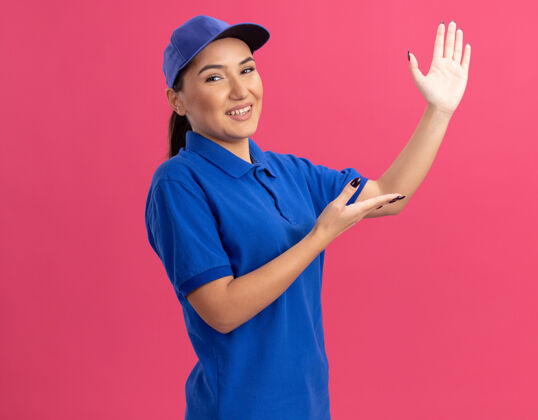 介绍身穿蓝色制服 头戴帽子的年轻送货员微笑着自信地站在粉红色的墙上 展示复印空间交货微笑帽子
