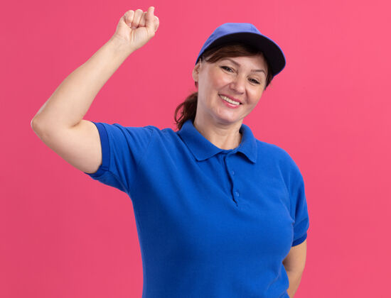 年龄身穿蓝色制服 头戴鸭舌帽的中年女送货员望着前面 愉快地微笑着 食指站在粉红色的墙上女人目录交货