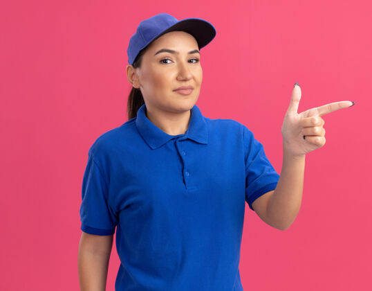 侧身身穿蓝色制服 头戴鸭舌帽的年轻女送货员微笑着自信地用食指指着站在粉色墙上的一侧帽子手指年轻