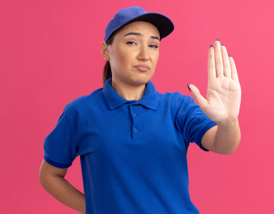 立场身穿蓝色制服 头戴鸭舌帽 表情严肃的年轻女送货员站在粉红色的墙上交货停止帽子